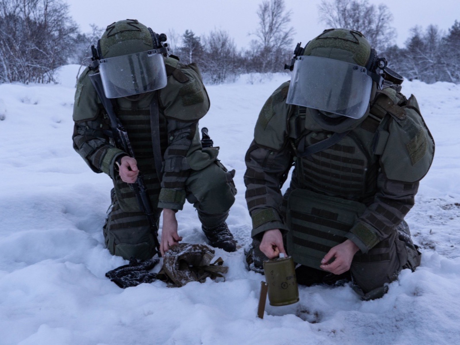 Два сапёра в защитных костюмах и шлемах с прозрачными забралами присели на снегу над учебным боеприпасом