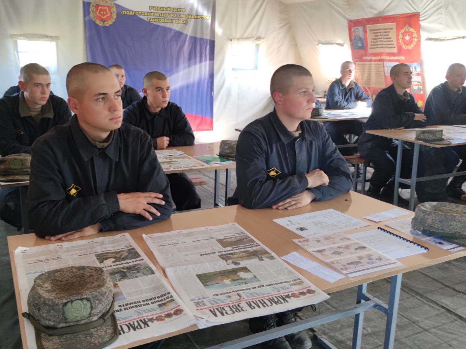 Занятие в палатке-классе учебного центра: новобранцы в тёмной форме сидят за столами с тетрадями и газетами, на стене флаг и стенд учебного центра